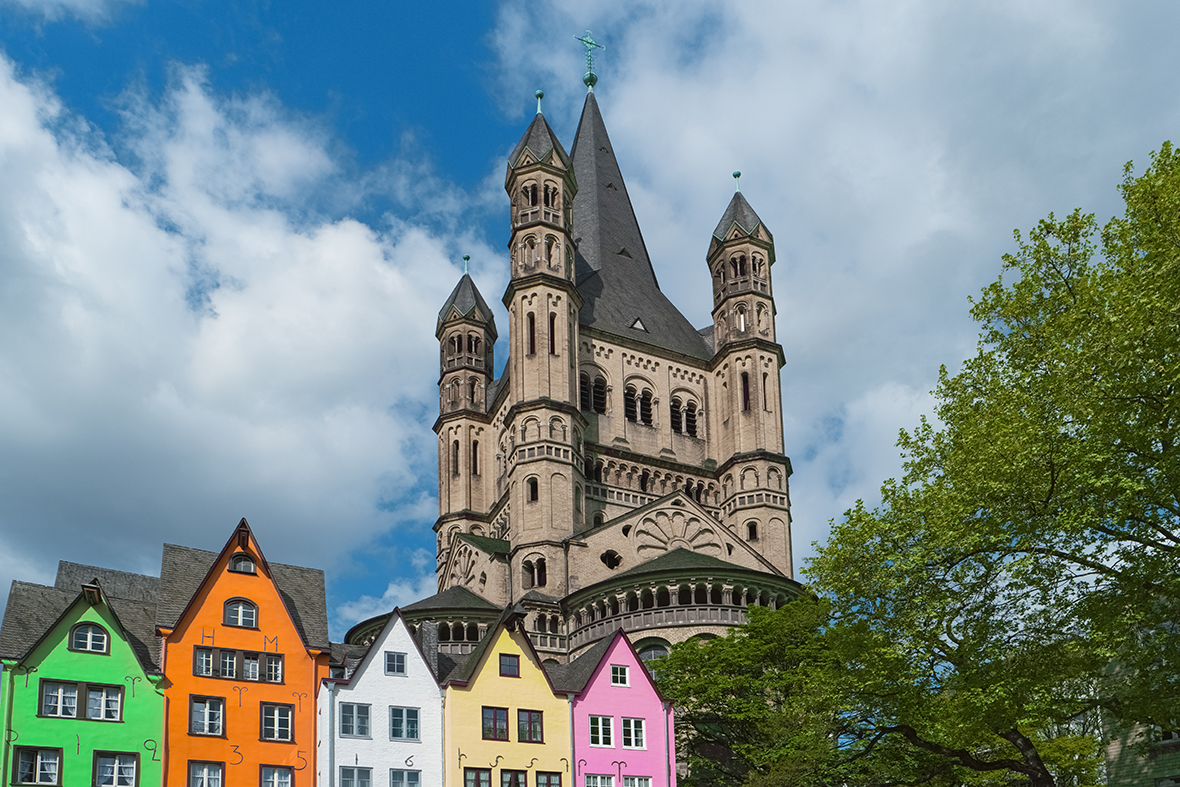 a row of colourful buildings in green, orange, pink, and white with a towering Gothic cathedral behind them and a blue sky above